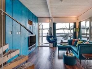 Een moderne woonkamer met een grote televisie, een blauwe zithoek, een hangstoel en een houten trap.