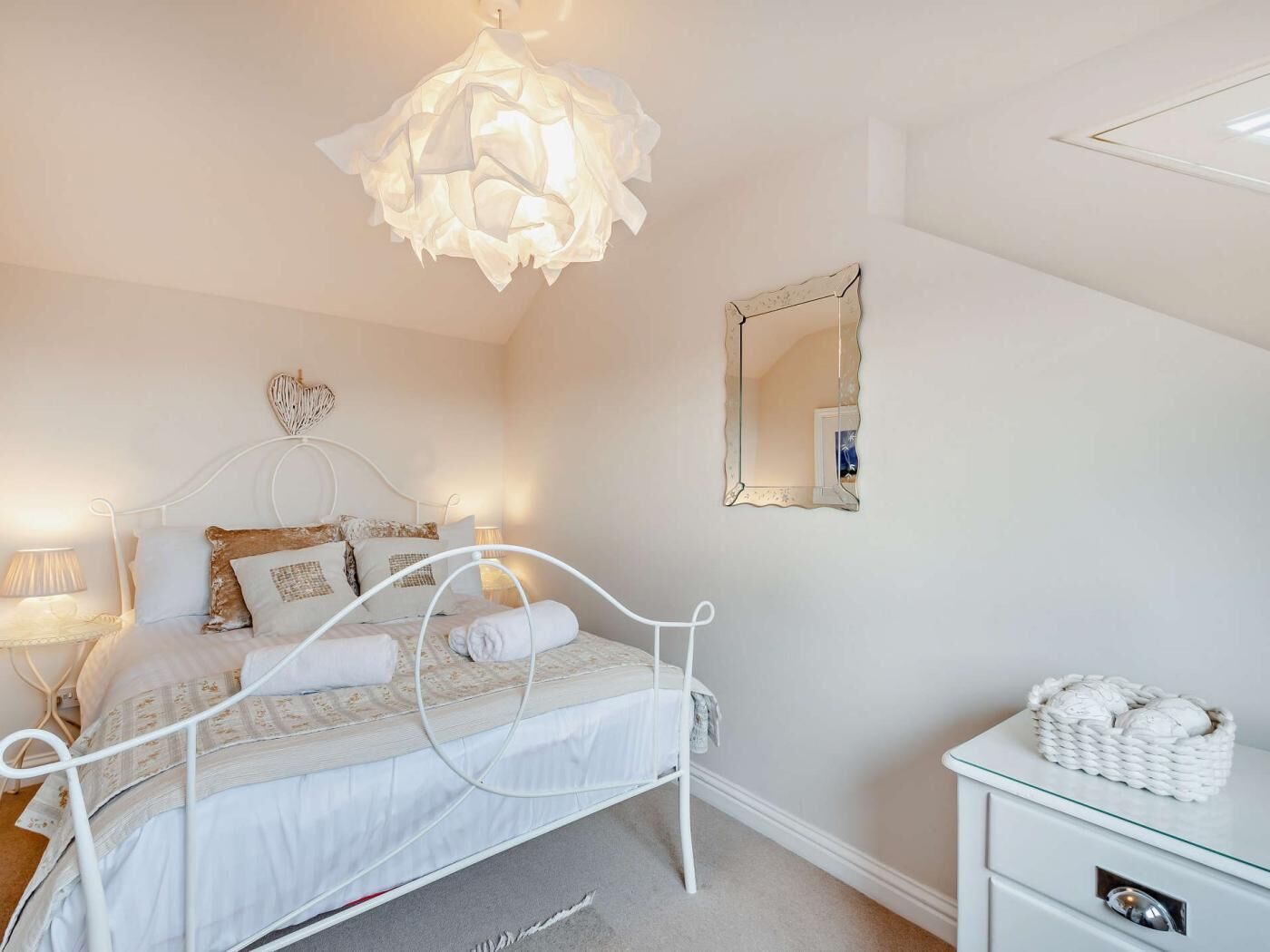 Een slaapkamer met een wit metalen bed, een bedlampje, een spiegel en een rieten mandje op een dressoir.