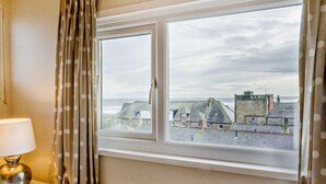 A room with a window overlooking a coastal town, a lamp on a bedside table, and a curtain with a polka dot pattern. - 3 Bed in Alnwick (oc-94013) (Alnmouth)