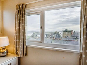 A room with a window overlooking a coastal town, a lamp on a bedside table, and a curtain with a polka dot pattern. - 3 Bed in Alnwick (oc-94013) (Alnmouth)