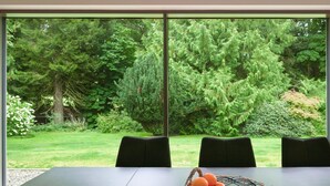 Uma mesa de jantar com uma cesta de frutas e quatro cadeiras em um quarto com grandes janelas com vista para um jardim exuberante.