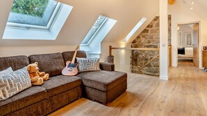 Um quarto no estilo loft com piso de madeira, um sofá seccional marrom, um violão e um urso de pelúcia. Há claraboias e uma parede de pedra à vista.