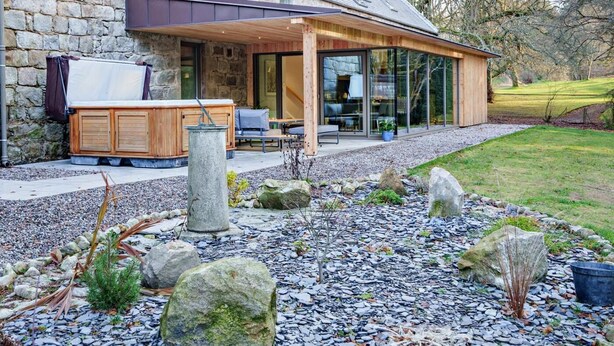 Uma casa moderna com fachada de pedra, um alpendre coberto e um jardim com pedras e plantas.