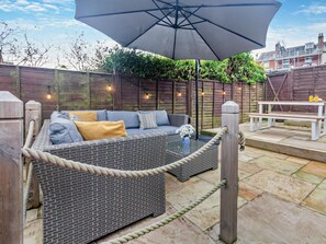 A wicker sofa with cushions, an umbrella, and a table with drinks, set in a patio area with a wooden fence and plants. - 3 Bed in Cowes (oc-93982) (Cowes)