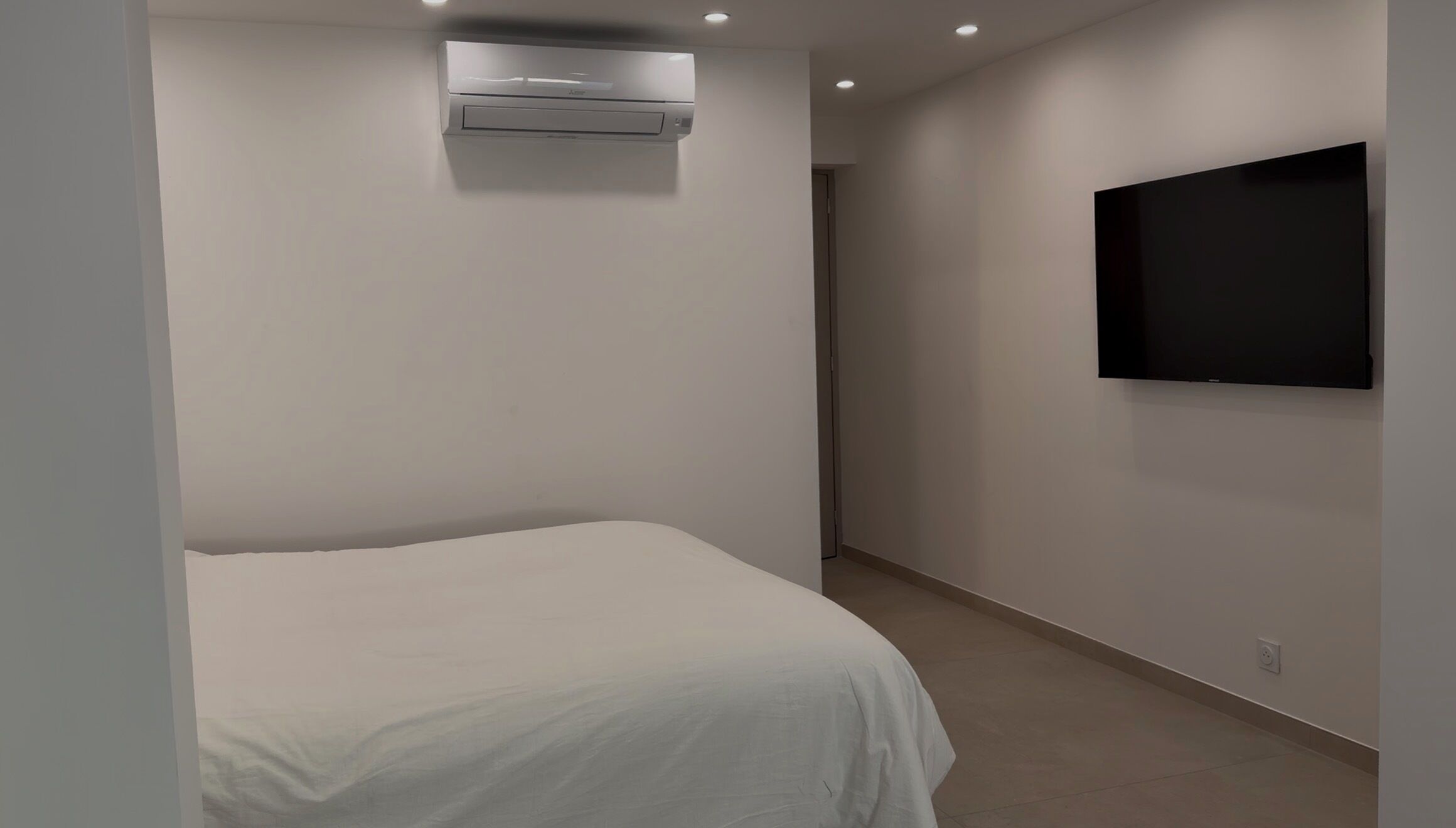 Una habitació amb un llit, un aparell d'aire condicionat, un televisor fixat a la paret i una porta que dona a una altra habitació.