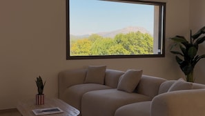 Un salon avec un canapé beige, une petite table, une plante en pot et une fenêtre donnant sur une vue sur les montagnes.
