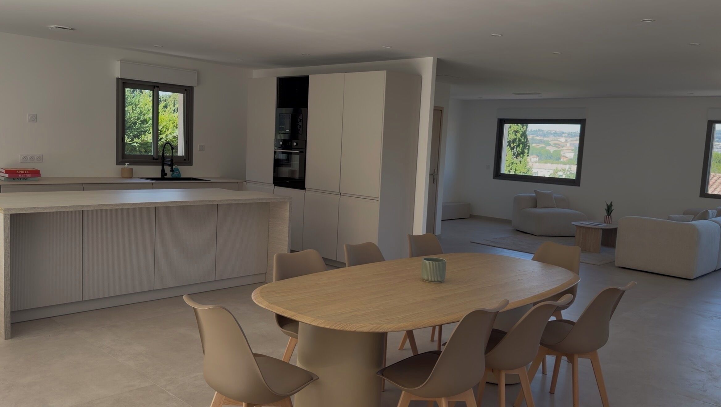 Eine moderne Küche mit einer zentralen Kochinsel, ein Essbereich mit rundem Tisch und Stühlen sowie ein Wohnbereich mit Sofa und Blick auf Grünflächen.