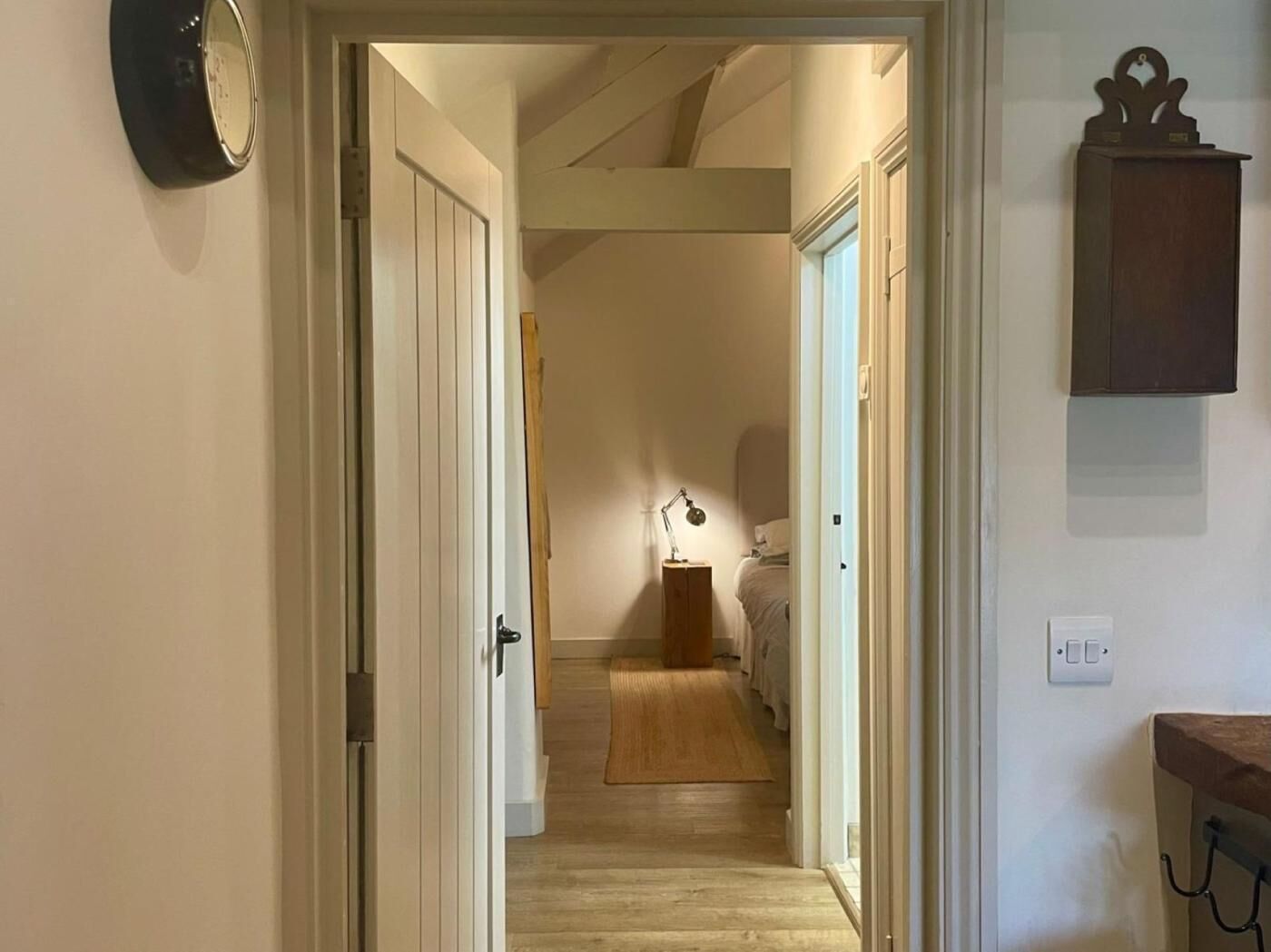 A hallway leading to a bedroom with a bed, a bedside table, and a lamp. There is a clock on the wall and a small cabinet on the right.
