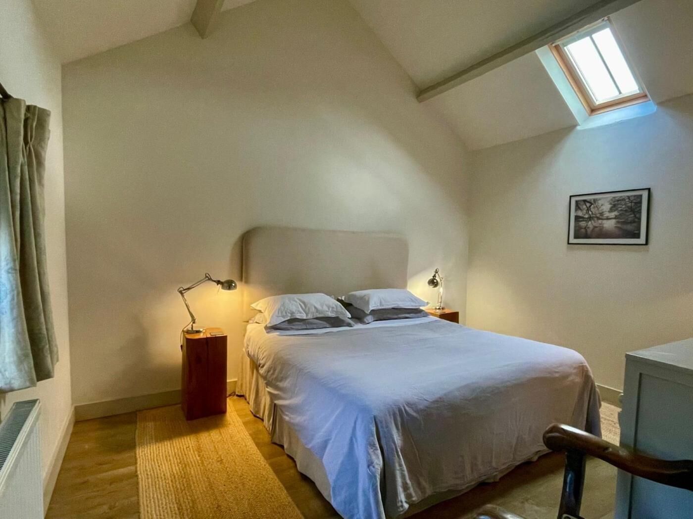 Ein Schlafzimmer mit einem Bett, zwei Nachttischlampen, einem Fenster und einem gerahmten Bild an der Wand.