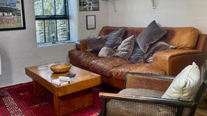 Un salon comprenant un canapé en cuir, une table basse en bois, un fauteuil en osier, un tapis à motifs rouges, une bibliothèque remplie de livres, un collage encadré et une fenêtre avec des stores.