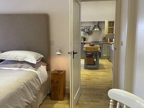 Ein Schlafzimmer mit einem Bett, einem Nachttisch und einem kleinen Tisch daneben. Ein Flur führt zu einer Küche mit Herd, Spüle und Schränken.