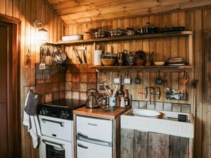 Eine Küche mit Holzwänden, einem Herd, einem Backofen sowie verschiedenen Küchenutensilien und Kochgeschirr auf Regalen.