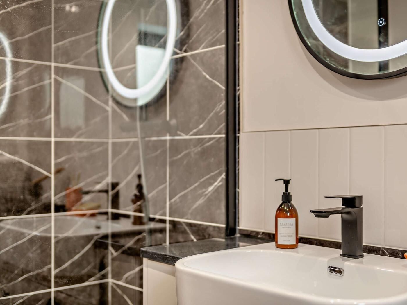 Une salle de bain avec un lavabo blanc, un robinet noir, un flacon de savon liquide et un miroir rond accroché au mur.
