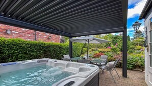 Ett bubbelbad under en pergola med en mur av tegelsten och en trädgård i bakgrunden.