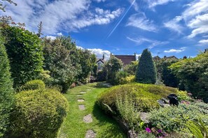 Ein Gartenpfad führt zu einem Haus mit rotem Dach, umgeben von üppiger Vegetation und verschiedenen Pflanzen.