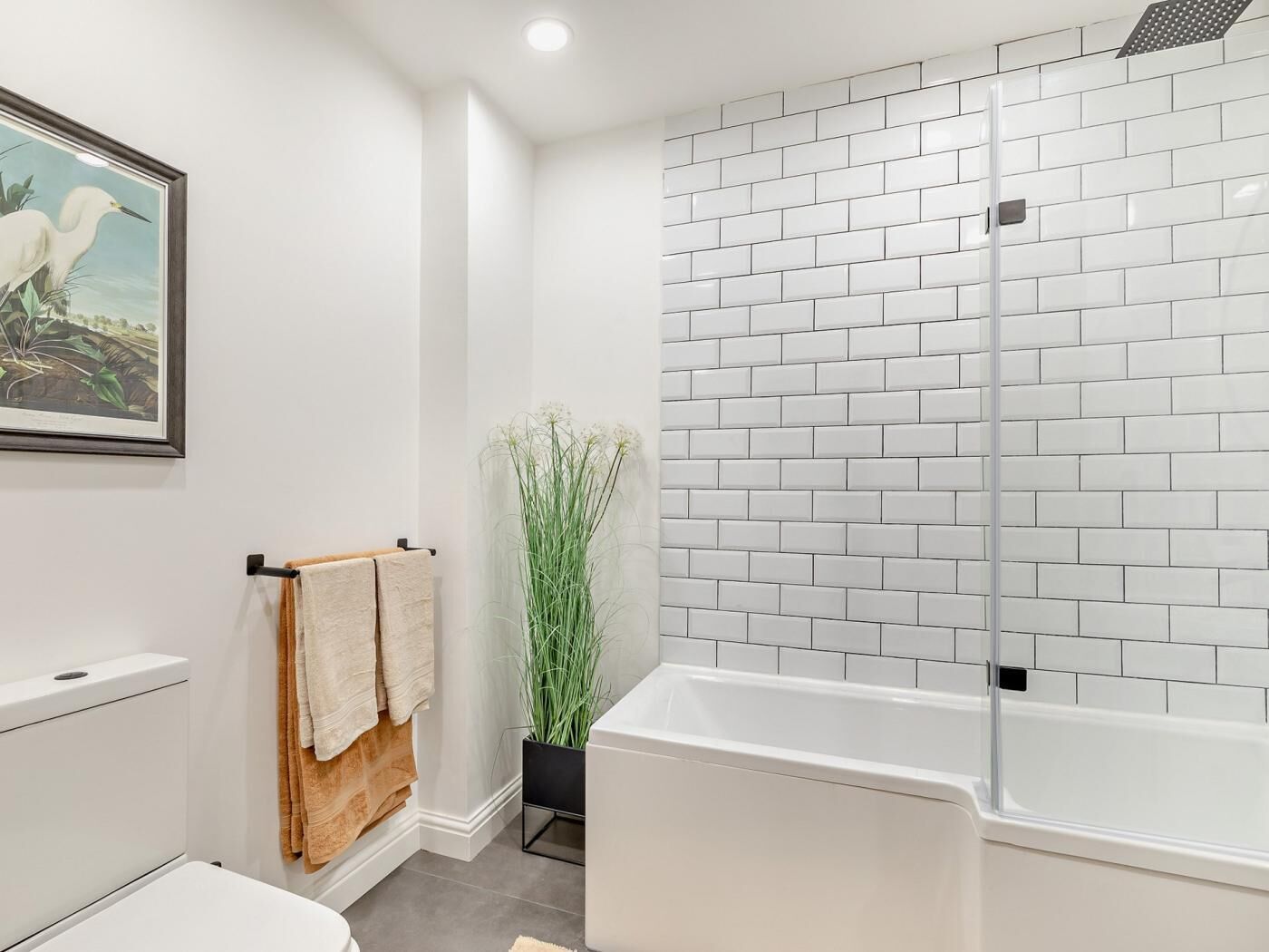 A modern bathroom with a white bathtub, a shower with white tiles, a toilet, a towel rack with towels, and a framed picture on the wall.