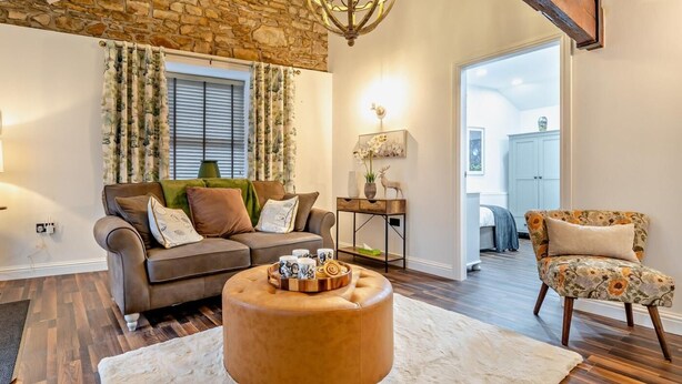 Una sala de estar con pared de piedra, techo de madera, un otoman redondo, un sofá, una mesita auxiliar y una silla.