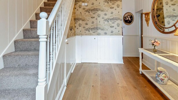 Una escalera con balaustres blancos, piso de madera, un candelabro, un espejo con marco dorado e ilustraciones náuticas en el papel tapiz.