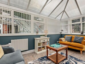 A conservatory with a white-framed glass roof, a grey sofa, a yellow sofa, a white cabinet with drawers, and a small table with a chessboard. - 4 Bed in Torquay (oc-94002) (Torquay)