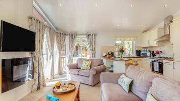 A living room with a TV, sofas, a coffee table with breakfast items, and a kitchen in the background.