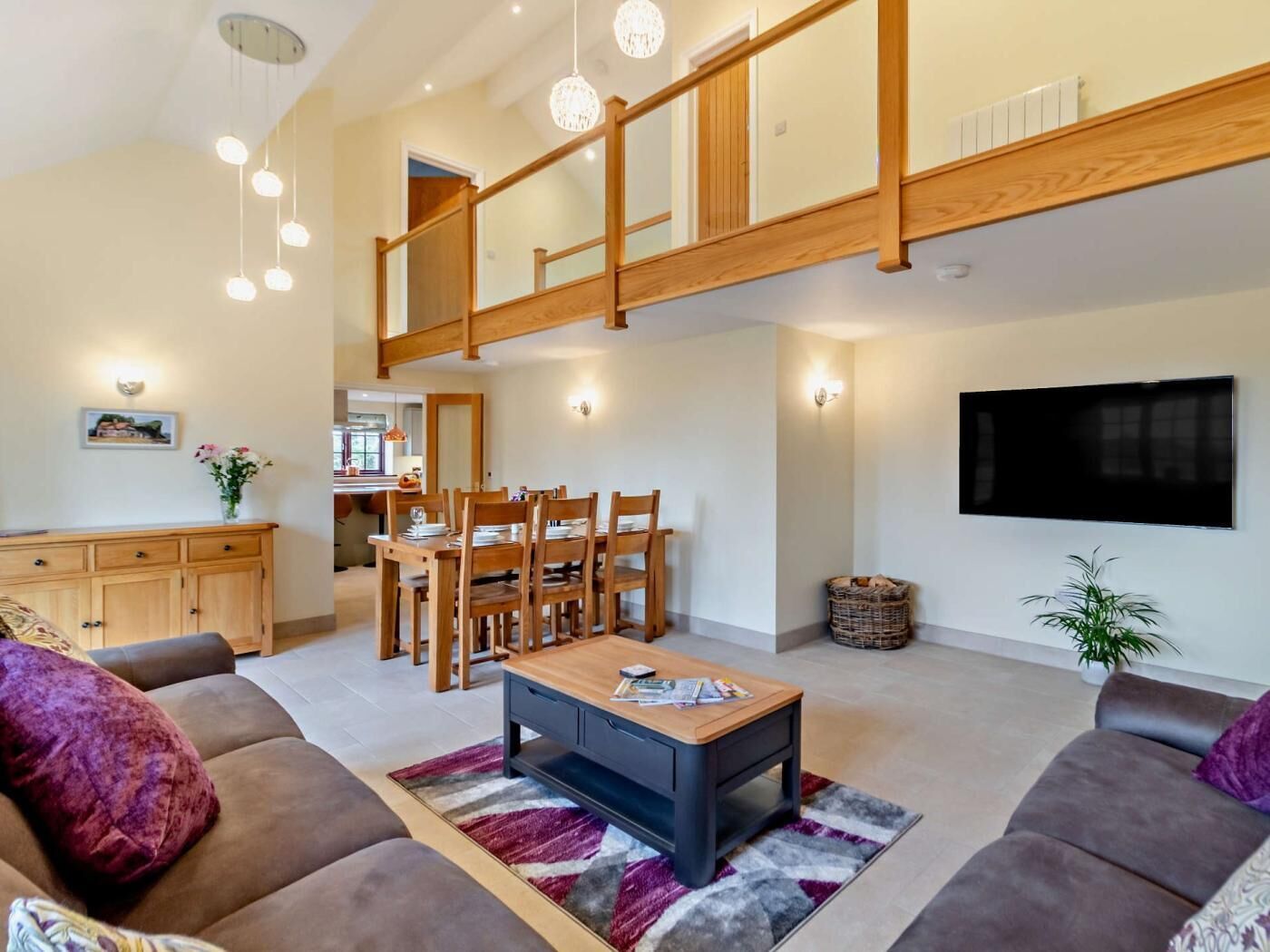 A spacious living area with a high ceiling, a dining table set for four, a sofa, a coffee table, and a television mounted on the wall.
