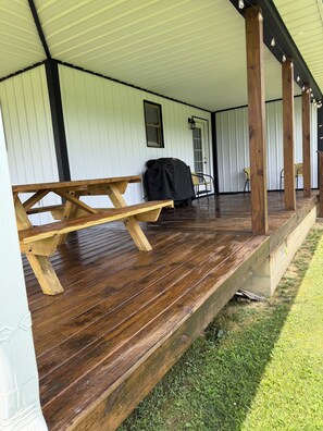 Een overdekt buitengebied met een houten picknicktafel, een barbecue en stoelen.