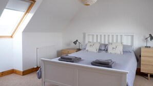 Un dormitorio con techo inclinado, tragaluz, cama con almohadas, mesita de noche, lámpara y radiador.