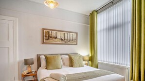 Une chambre d’hôtel bien rangée, avec un lit, une table de chevet, une lampe, un tableau encadré au mur et une fenêtre avec des rideaux.