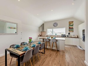 A modern kitchen with a central island, a dining area with a wooden table and chairs, and a large window providing natural light. - 3 Bed in Westward Ho! (oc-78075) (Westward Ho!)