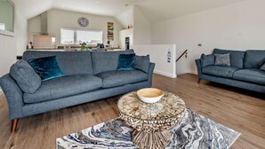 Ein geräumiger Wohnbereich mit einem grauen Sofa, einem runden Couchtisch mit einer Schale und einer Küche im Hintergrund.