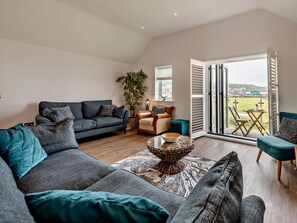 A modern living room with a grey sofa, a coffee table, and a view through a glass door to an outdoor area. - 3 Bed in Westward Ho! (oc-78075) (Westward Ho!)