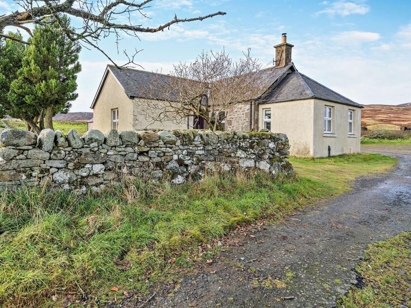 A stone house with a stone wall, a gravel driveway, and a grassy area with trees.