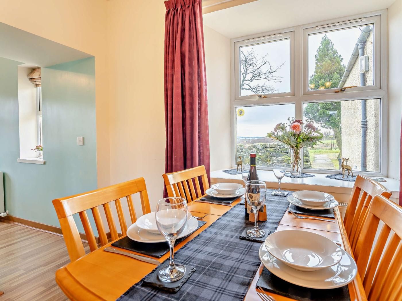 A dining room with a table set for four, wooden chairs, a window with a view of greenery, and a red curtain.