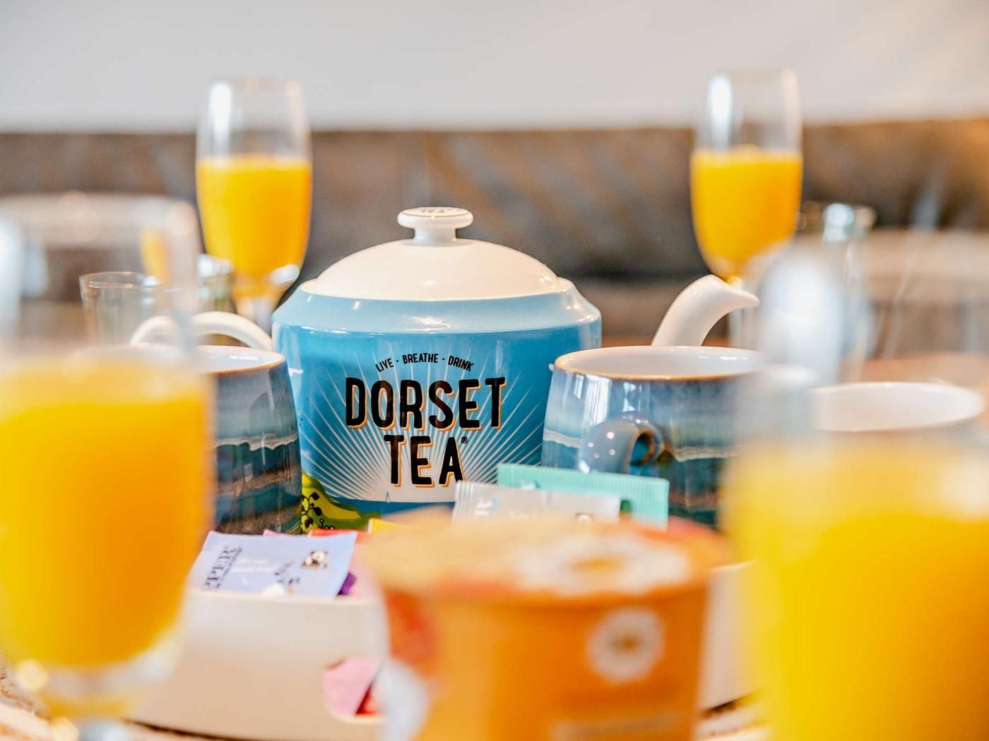 Een gedekte tafel met een blauwe theepot met de tekst 'Dorset Tea' en verschillende glazen sinaasappelsap.