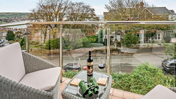 Un balcon agrémenté de meubles en osier, d’une table en verre et d’une bouteille de vin.