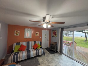 Une pièce avec un canapé à rayures, un ventilateur de plafond, une porte et une vue sur une terrasse et l’eau à l’extérieur.