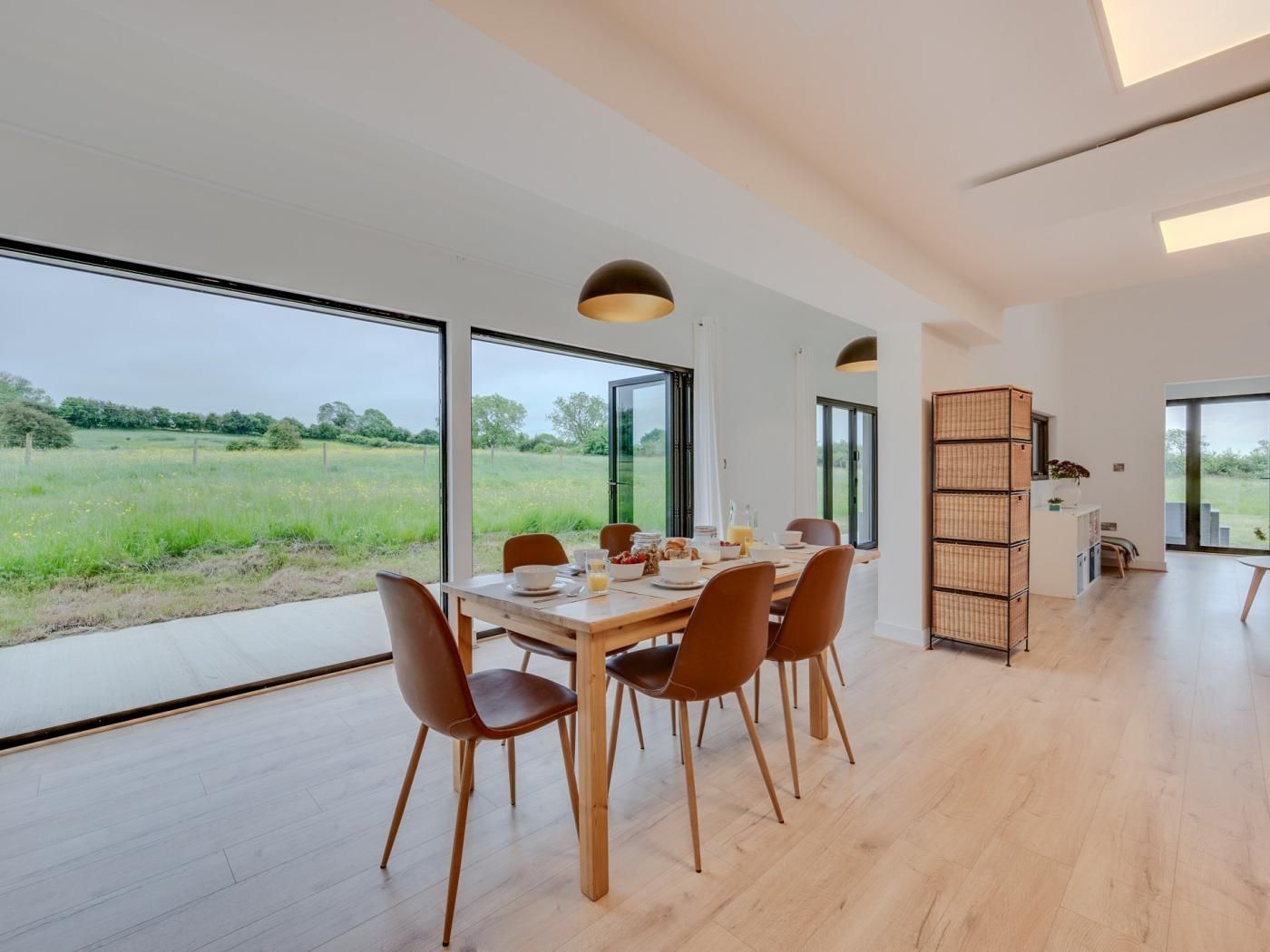 A modern dining area with a large table and chairs, large windows offering a view of a green field, and a built-in shelving unit.