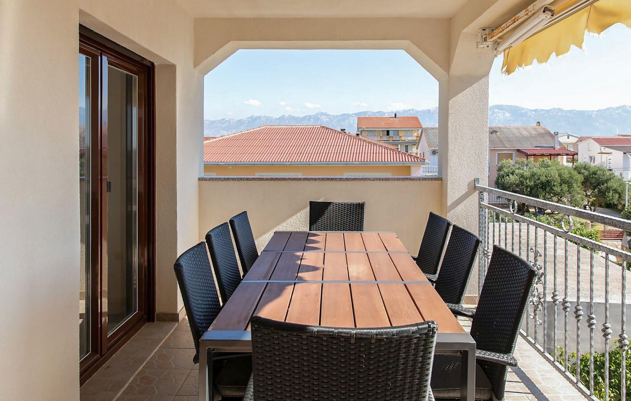 Un balcon avec une table et des chaises en bois, une vue sur les bâtiments et les montagnes, et un auvent jaune.