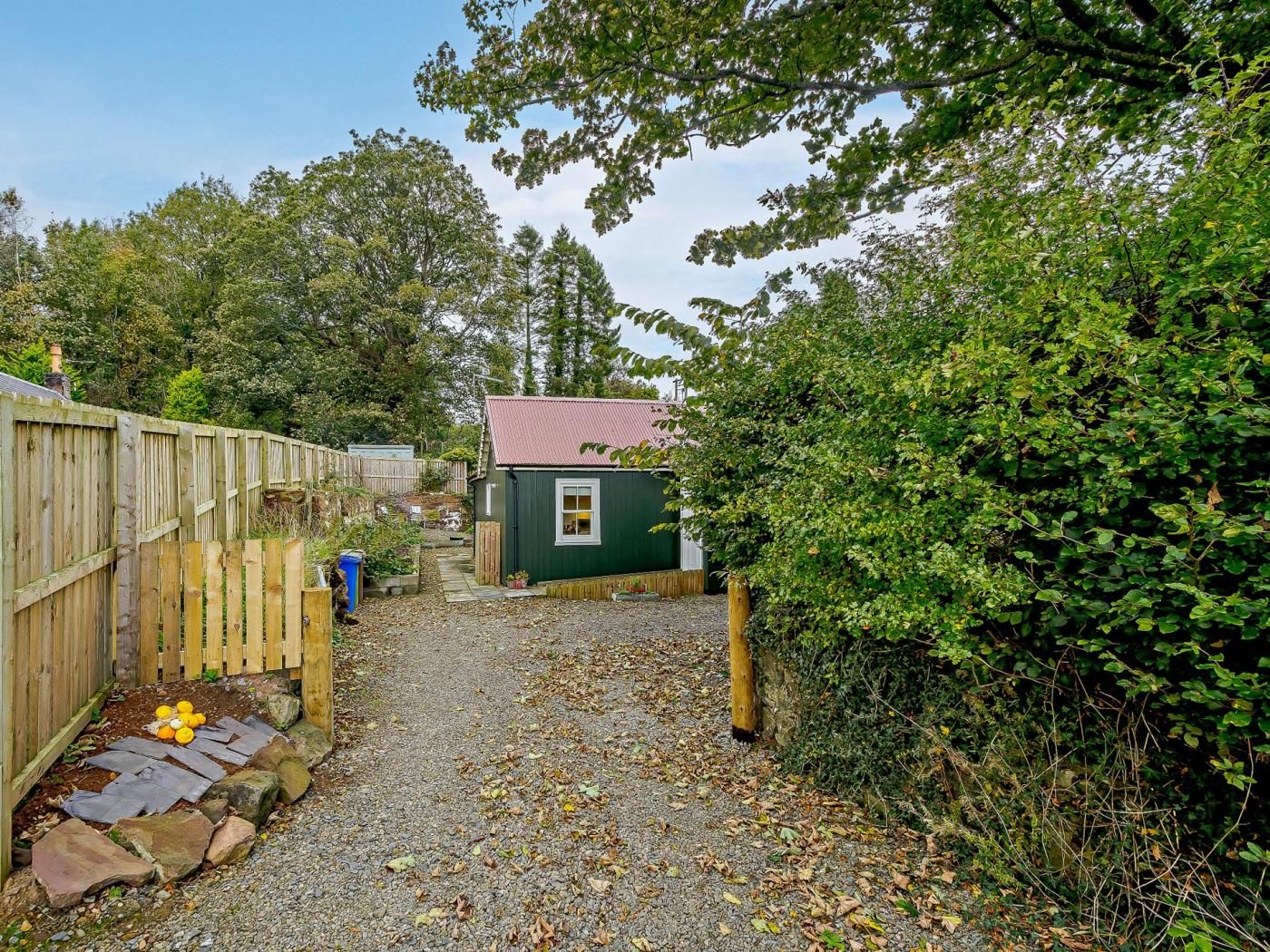 Ein Kiesweg führt zu einer kleinen grünen Gartenhütte mit rotem Dach, umgeben von einem Holzzaun und dichtem Grün.