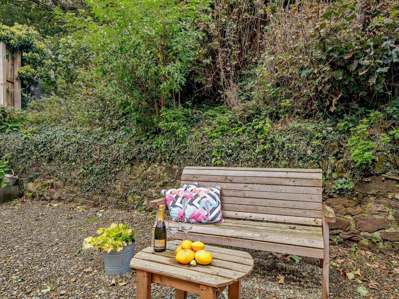 Un banco de madera con cojín, una botella de champán, una mesita con planta en maceta y limones, y un muro de piedra al fondo.