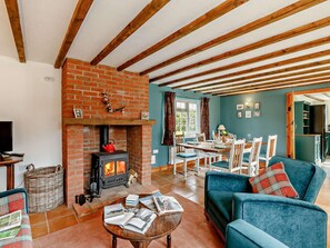 A cozy living room with a brick fireplace, wooden beams, a dining area, and a kitchen visible through an open door.