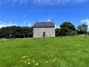 Ein aus Stein gebautes Haus mit dunklem Dach, umgeben von einem grasbewachsen Feld und Bäumen.