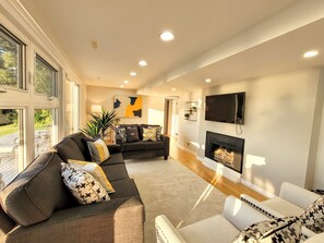 A spacious living room with a fireplace, a television mounted on the wall, and a large window providing natural light. - Relaxing Waterfront Cottage (Prince Edward)