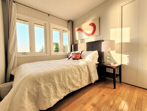 Ein Schlafzimmer mit einem Bett, einem Nachttisch, einer Lampe, einem Teddybären und einem Fenster mit Vorhängen.