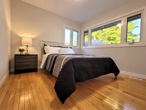 A bedroom with a bed, a nightstand with a lamp, and a window with a view of trees. - Relaxing Waterfront Cottage (Prince Edward)