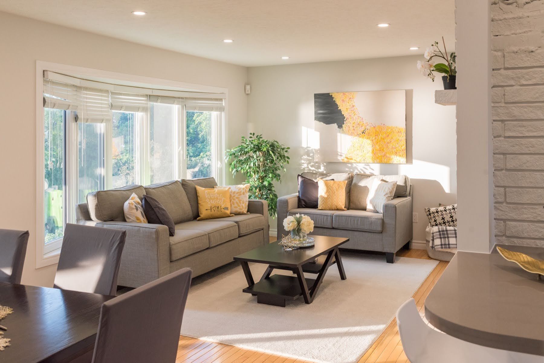 A living room with a sofa, coffee table, and chairs, a large window with blinds, and a wall-mounted artwork.