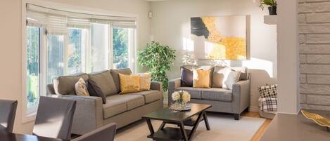 A living room with a sofa, coffee table, and chairs, a large window with blinds, and a wall-mounted artwork.