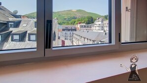 Udsigt gennem et vindue af en bygning med flere etager, hvor 'HOTEL GLORIA' er synligt, og en grøn bakke i baggrunden.