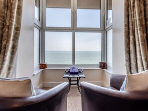 Ein Zimmer mit großem Fenster, zwei Ledersesseln, einem kleinen Tisch mit einem Glas und Blick auf das Meer.