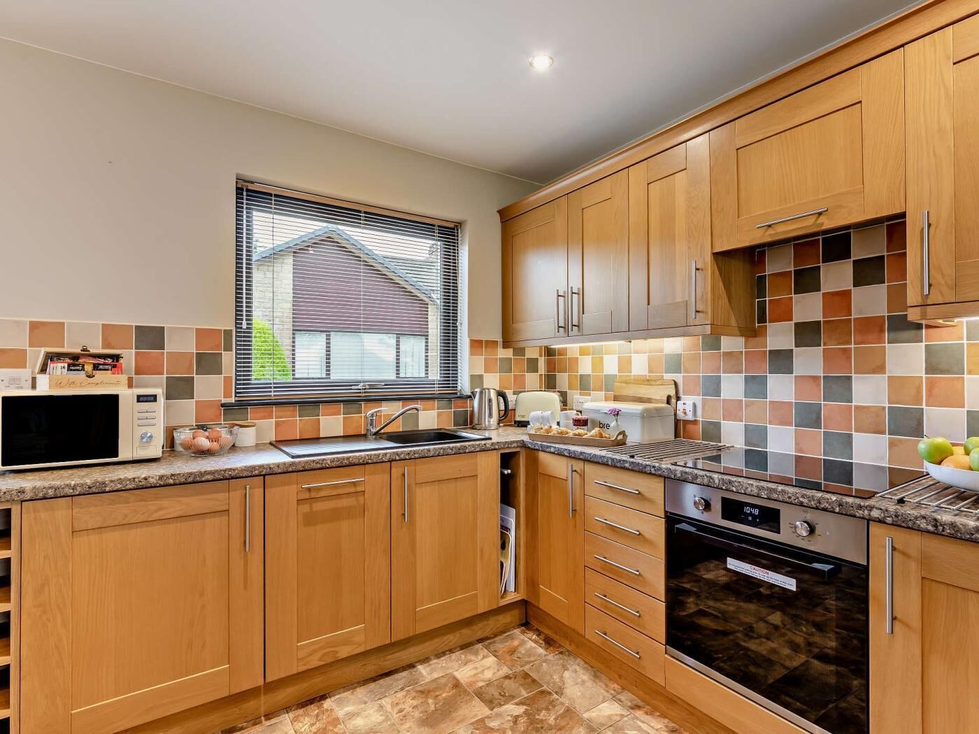 A modern kitchen with wooden cabinets, a built-in oven, a microwave, and a window with blinds.
